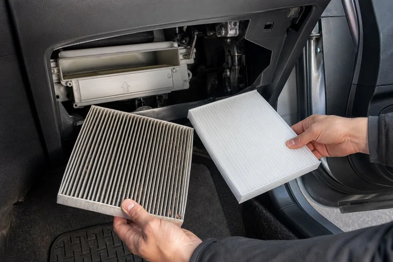 Hands removing a pleated cabin air filter behind an unbranded compact-car glovebox with old and clean filters visible